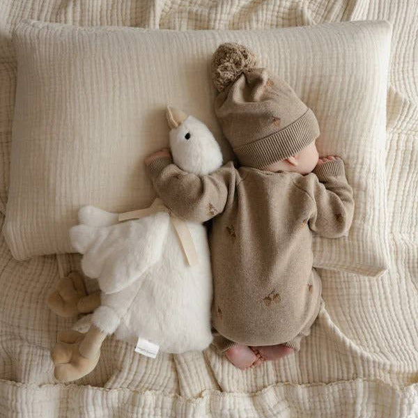 Baby in brown outfit and hat lying on a textured blanket with a plush toy.