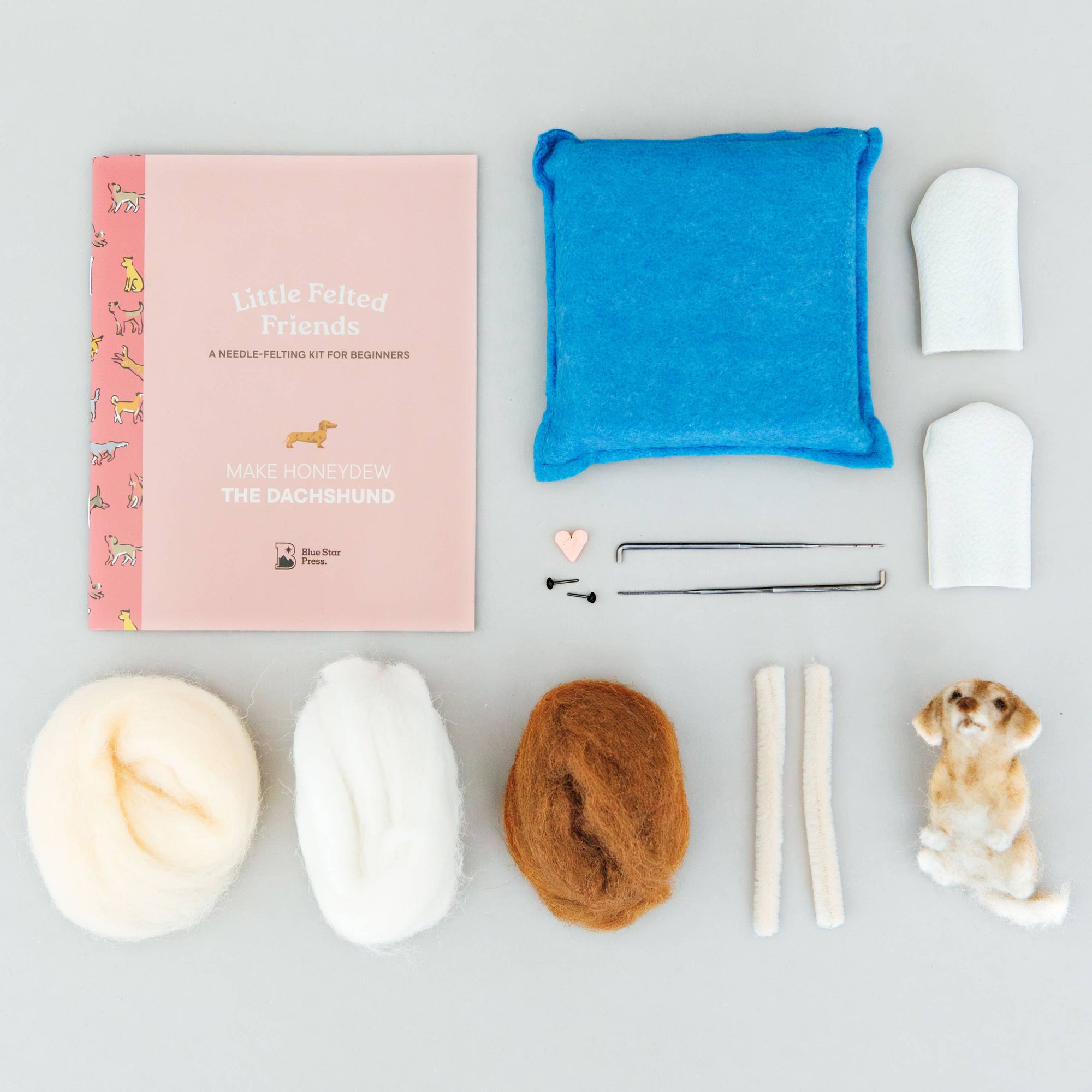 A flat-lay view of a needlework kit on a light gray background, including a pink patterned booklet, a blue felt pad, white and brown wool, metal needles, and a small dog-shaped piece of wool.