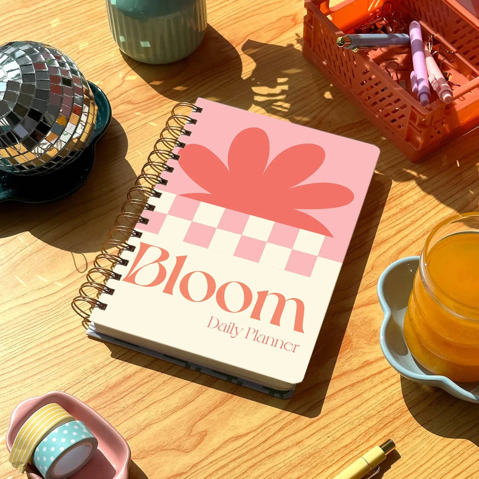 Notebook titled 'Bloom Daily Planner' on a wooden table with a cup of tea and decorative items.