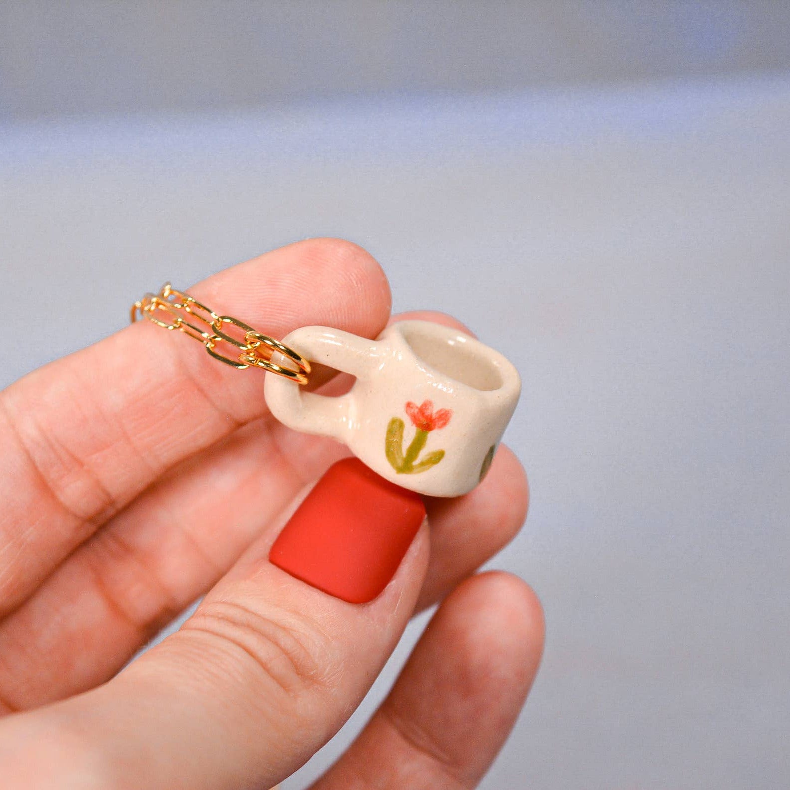 Small ceramic mug charm with a red base and gold chain held in a hand against a neutral background