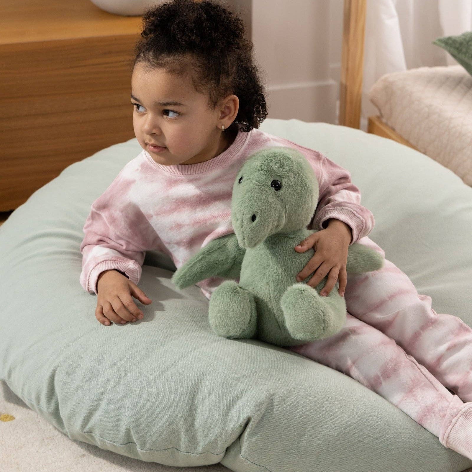 Child sitting on a large round pillow holding a green plush toy in a cozy room.