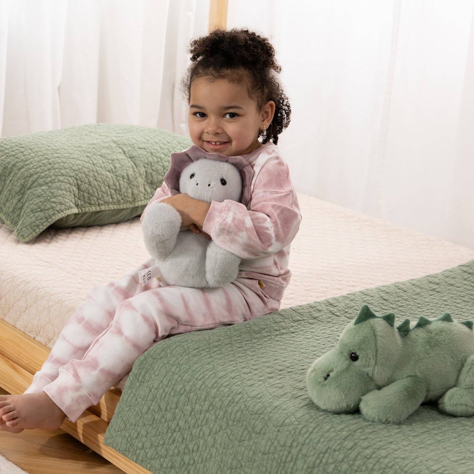 Child sitting on a bed with green bedding and holding a plush toy, surrounded by dinosaur toys.