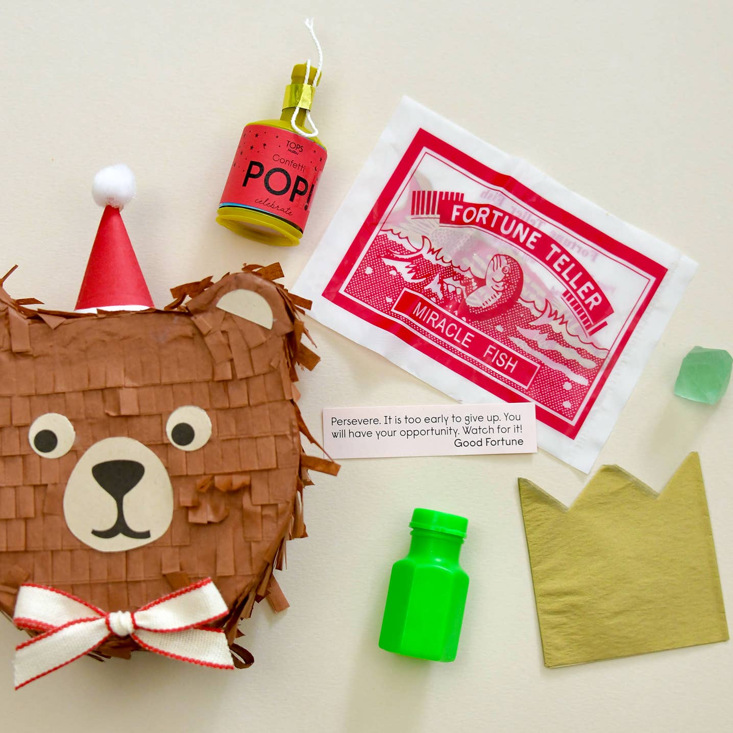 A flat-lay of a fortune-telling kit on a light beige background, featuring a brown bear pièna with a party hat and bow, a small bottle of 'POP' candy, a 'Fortune Teller Miracle Fish' card, a green bottle, gold paper, and a small green crystal.