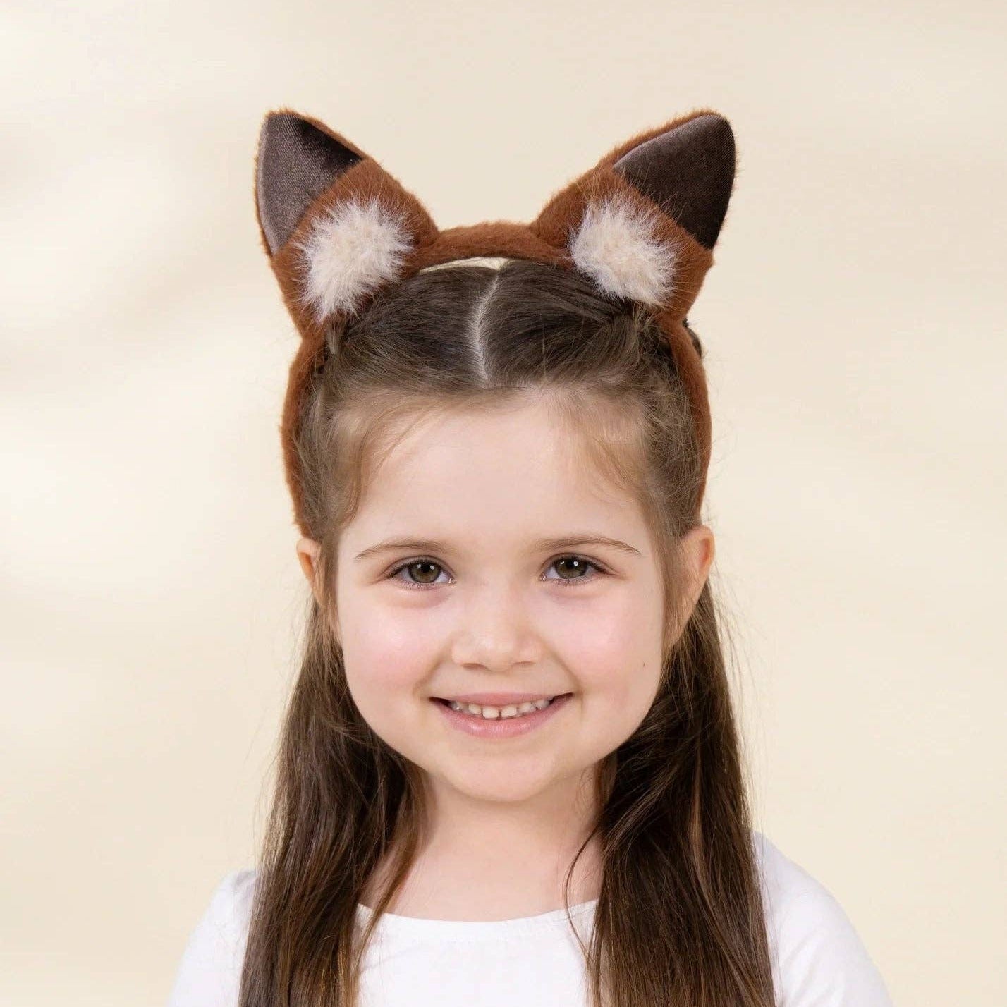 A young girl with long brown hair wearing a brown and white faux fur fox ear headband, photographed against a solid light beige background.