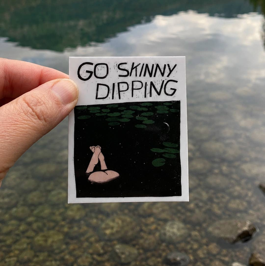 Hand holding a sticker with 'Go Skinny Dipping' text over a lake background