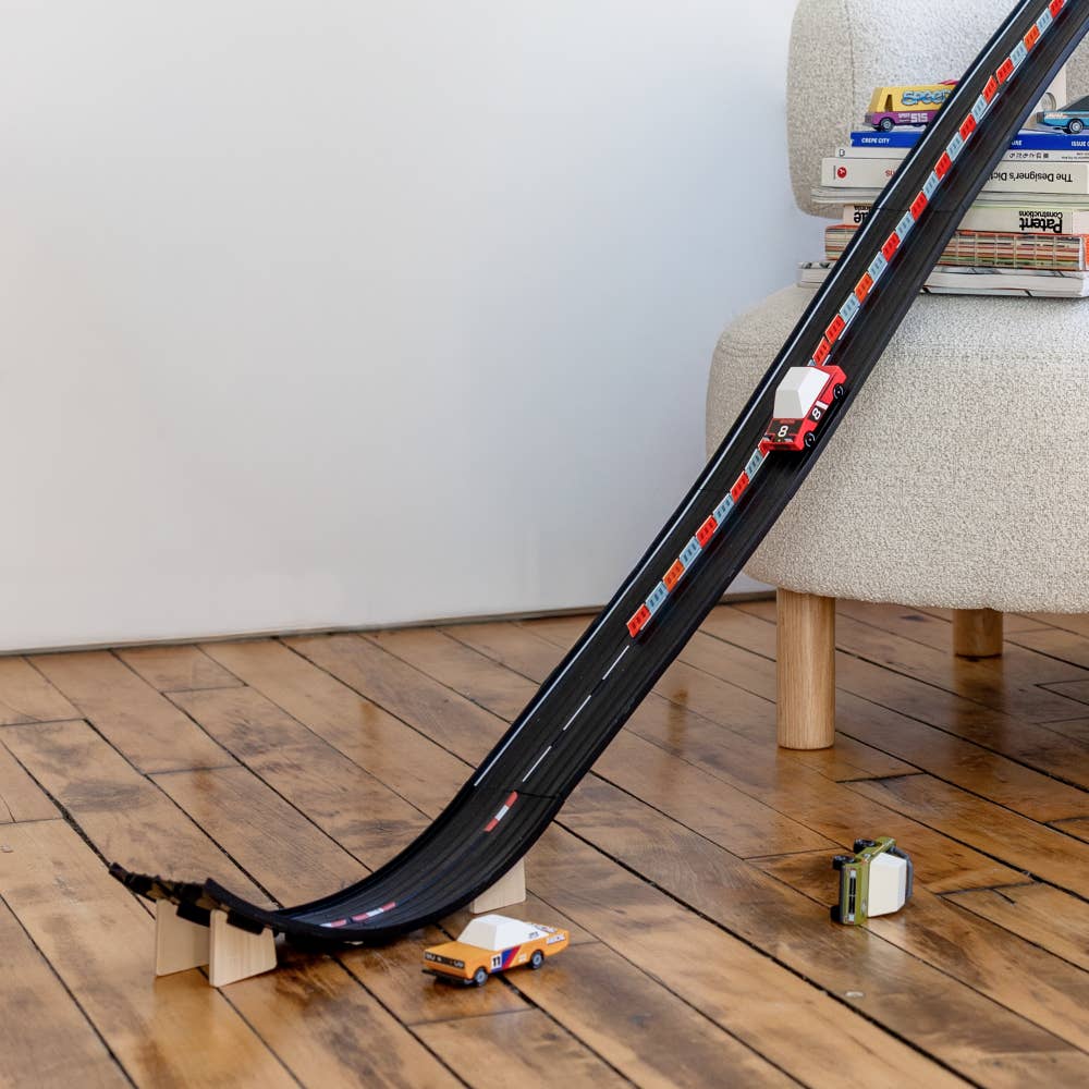 Toy car track set on a wooden floor with books stacked on a chair in the background.