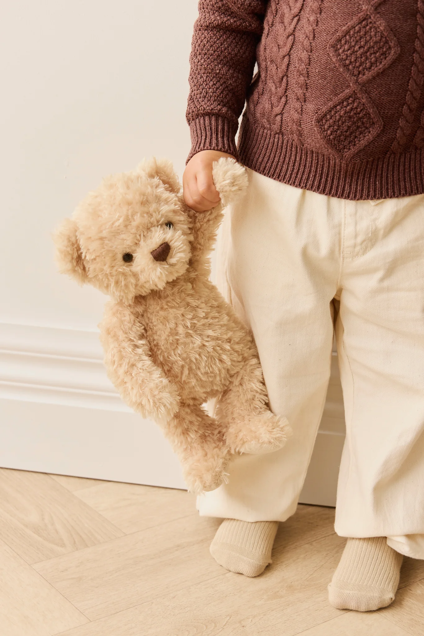 Person holding a teddy bear wearing a brown sweater and white pants.