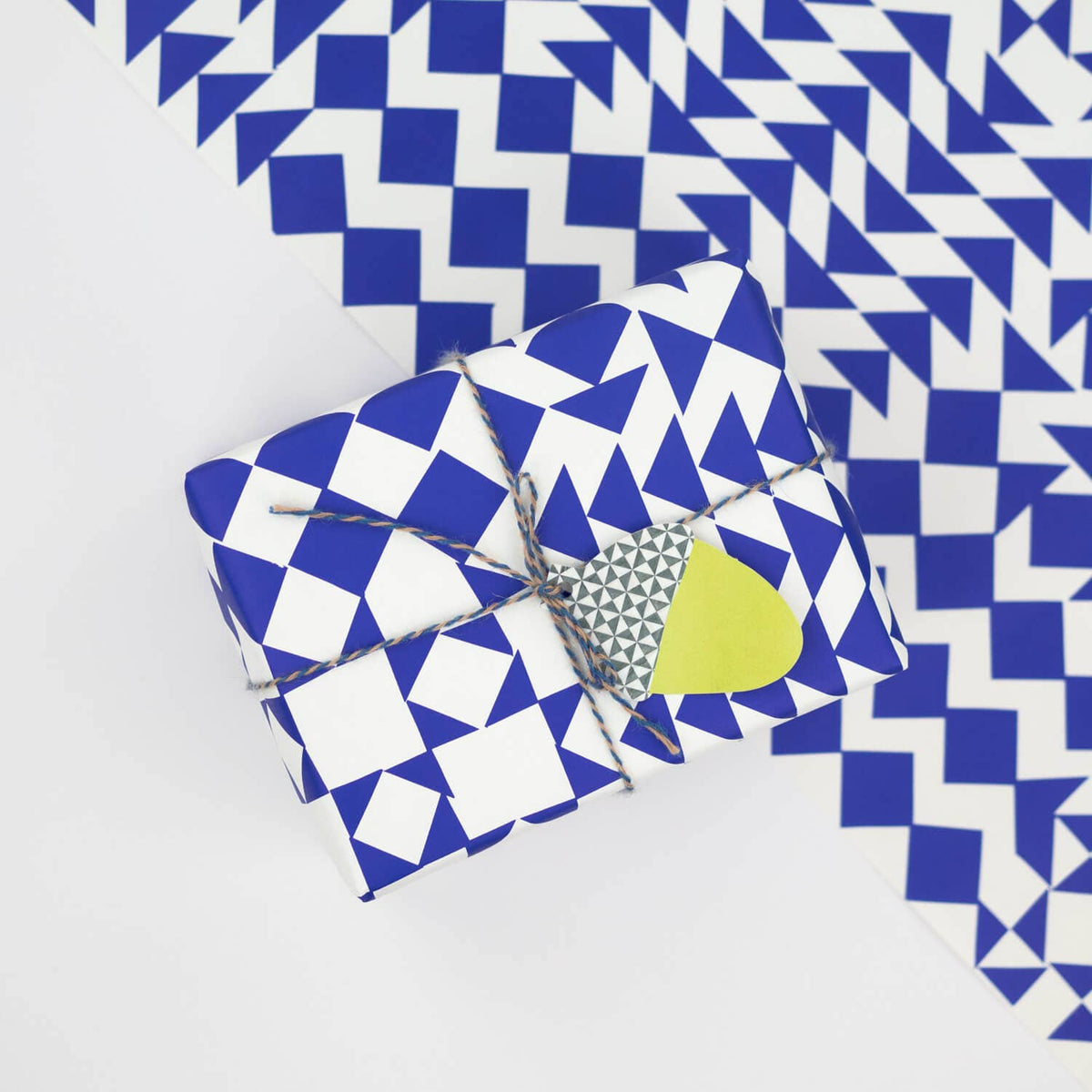 A wrapped gift box and matching wrapping paper featuring a bold blue and white geometric pattern of diamonds and triangles, tied with twine and decorated with a small yellow heart tag.