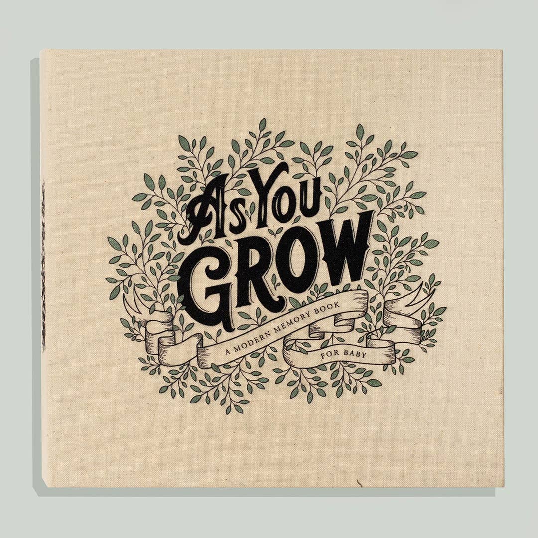 A square canvas-style baby memory book cover with the phrase “As You GROW” in large black lettering, surrounded by illustrated green leaves and vines on a warm cream background. Smaller text reads “A MODERN MEMORY BOOK FOR BABY.