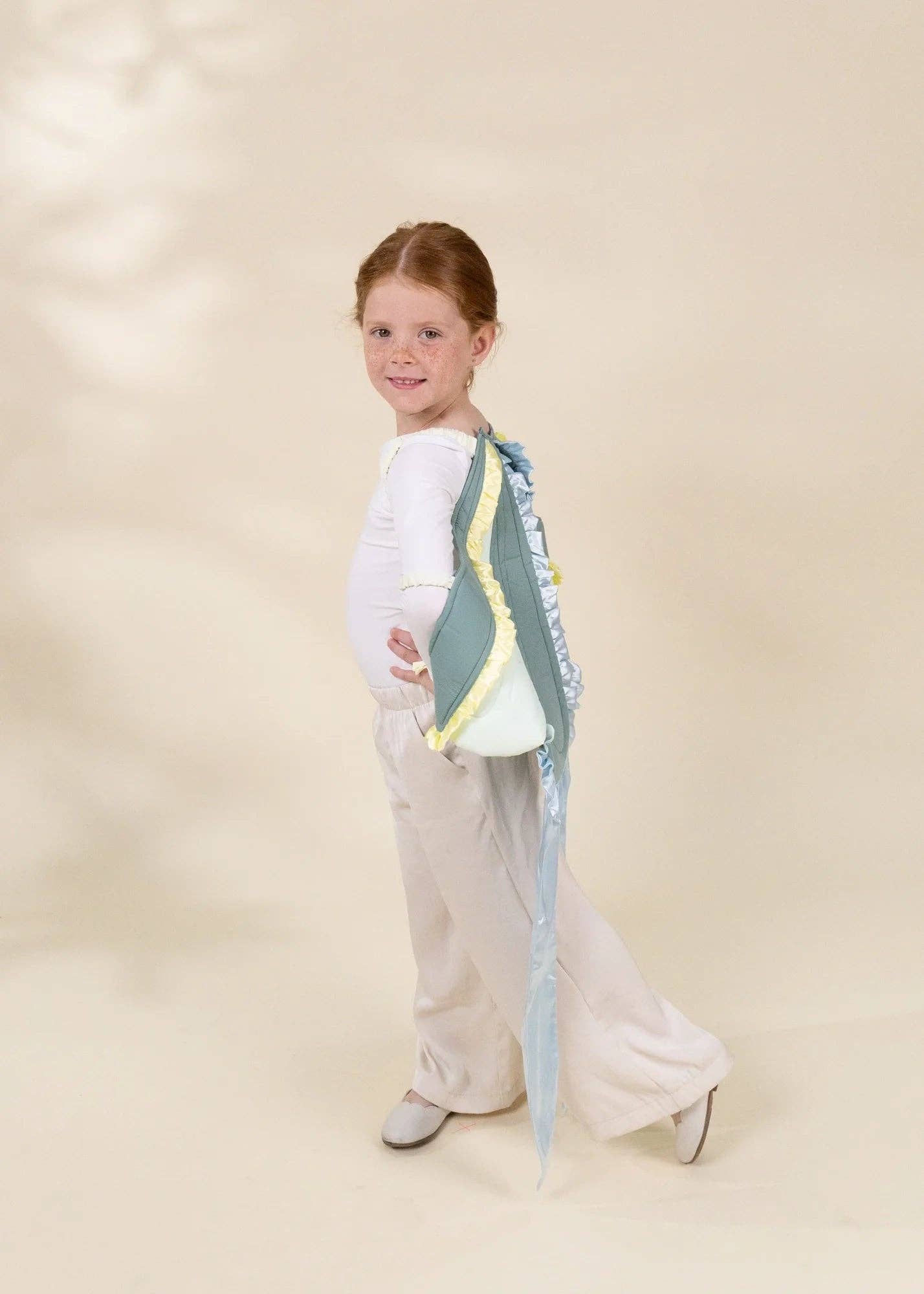 A young girl with red hair tied back is shown from the back wearing a white long-sleeve top, cream wide-leg pants, and gray shoes. She is carrying a light blue and white shoulder bag with yellow trim and a long blue ribbon.