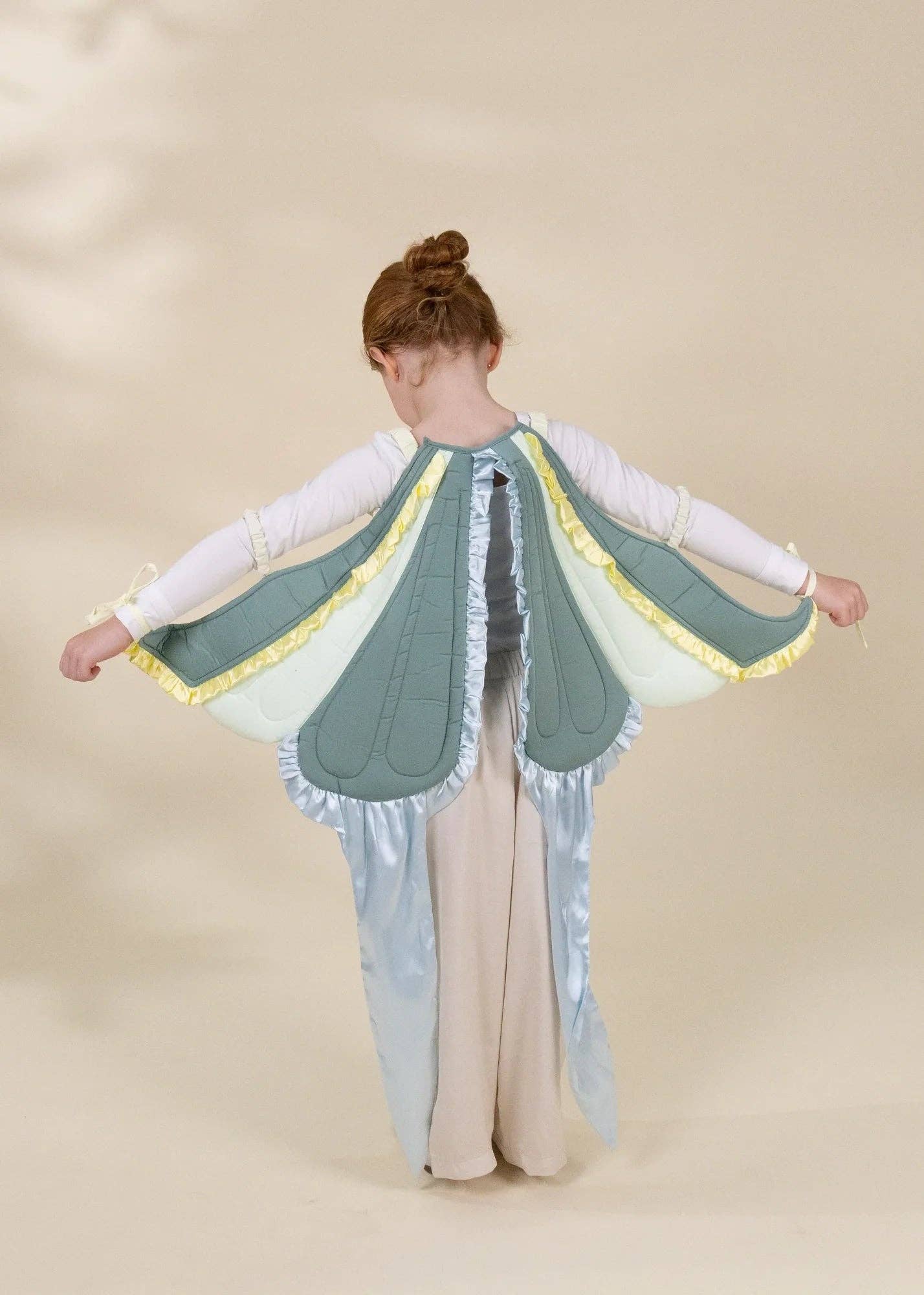 A child wearing a fairy or winged costume, shown from the back with arms outstretched. The costume features pale blue wings with yellow trim, a white long-sleeve top, and a beige skirt, set against a plain beige studio background.