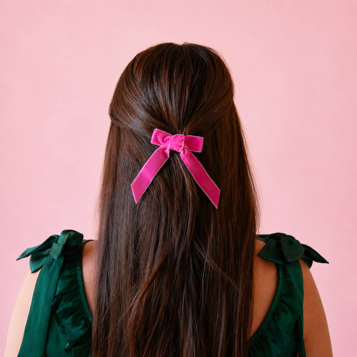 A person viewed from the back with long dark brown hair tied at the back with a bright pink velvet bow. The person is wearing a dark green sleeveless top with bow details on the shoulders, set against a solid light pink background.
