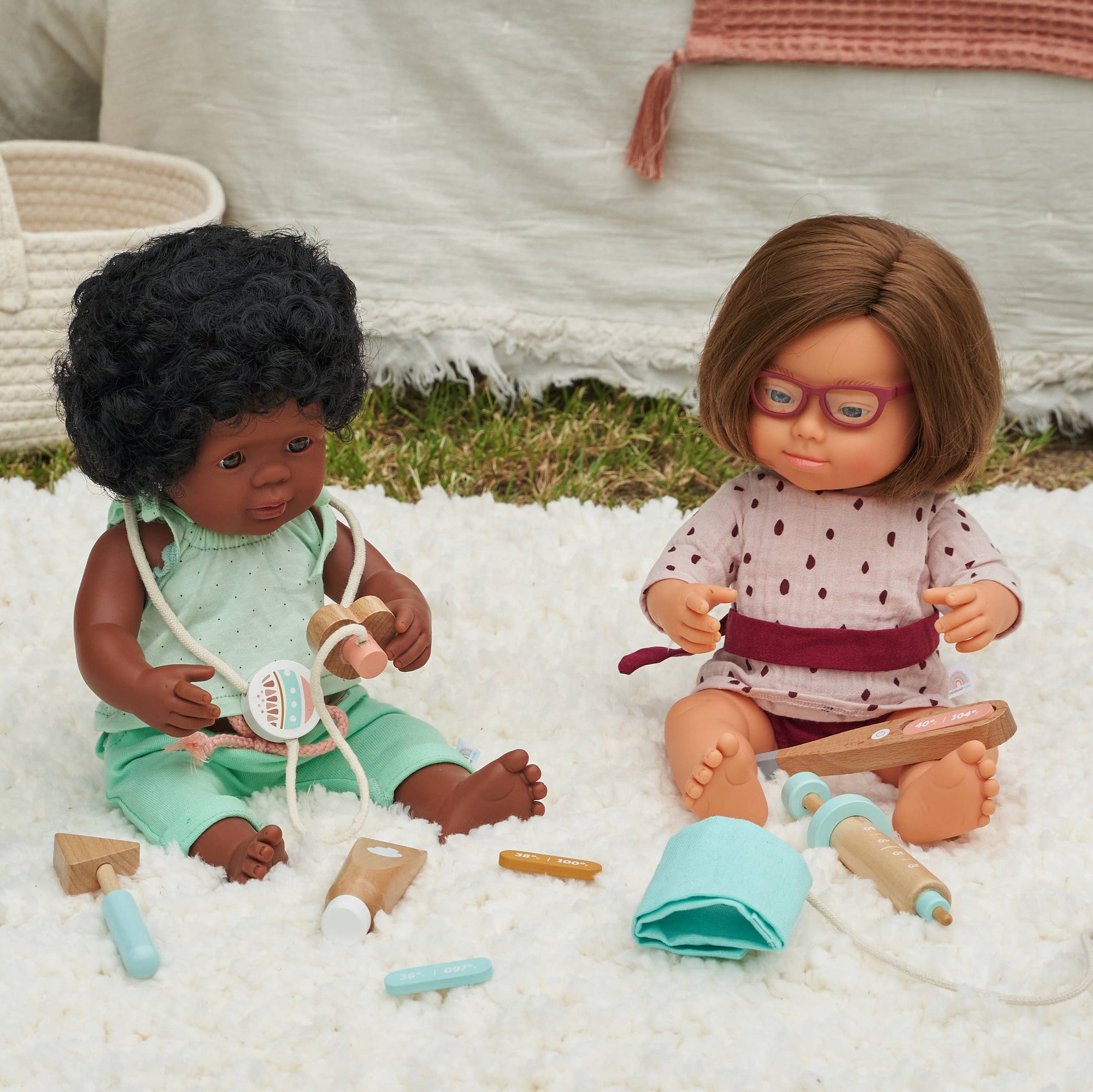 Two dolls sitting on a white surface with toys around them, one doll wearing glasses.