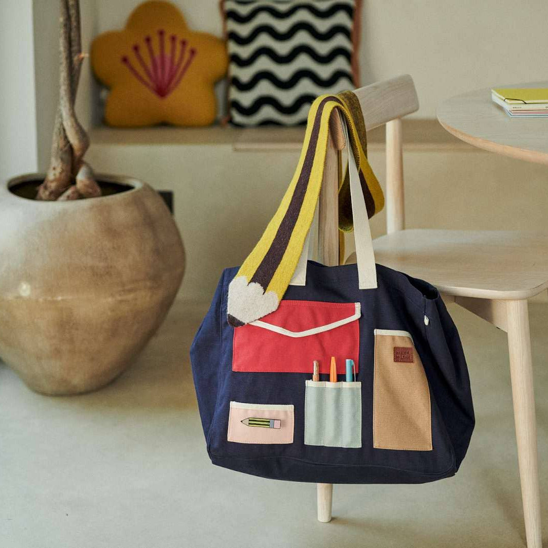 A navy tote bag with yellow and white shoulder straps hangs from a light wood chair in a bright room. The front of the bag has multiple utility pockets in red, beige, and light pink, with pens visible in a pen holder pocket.