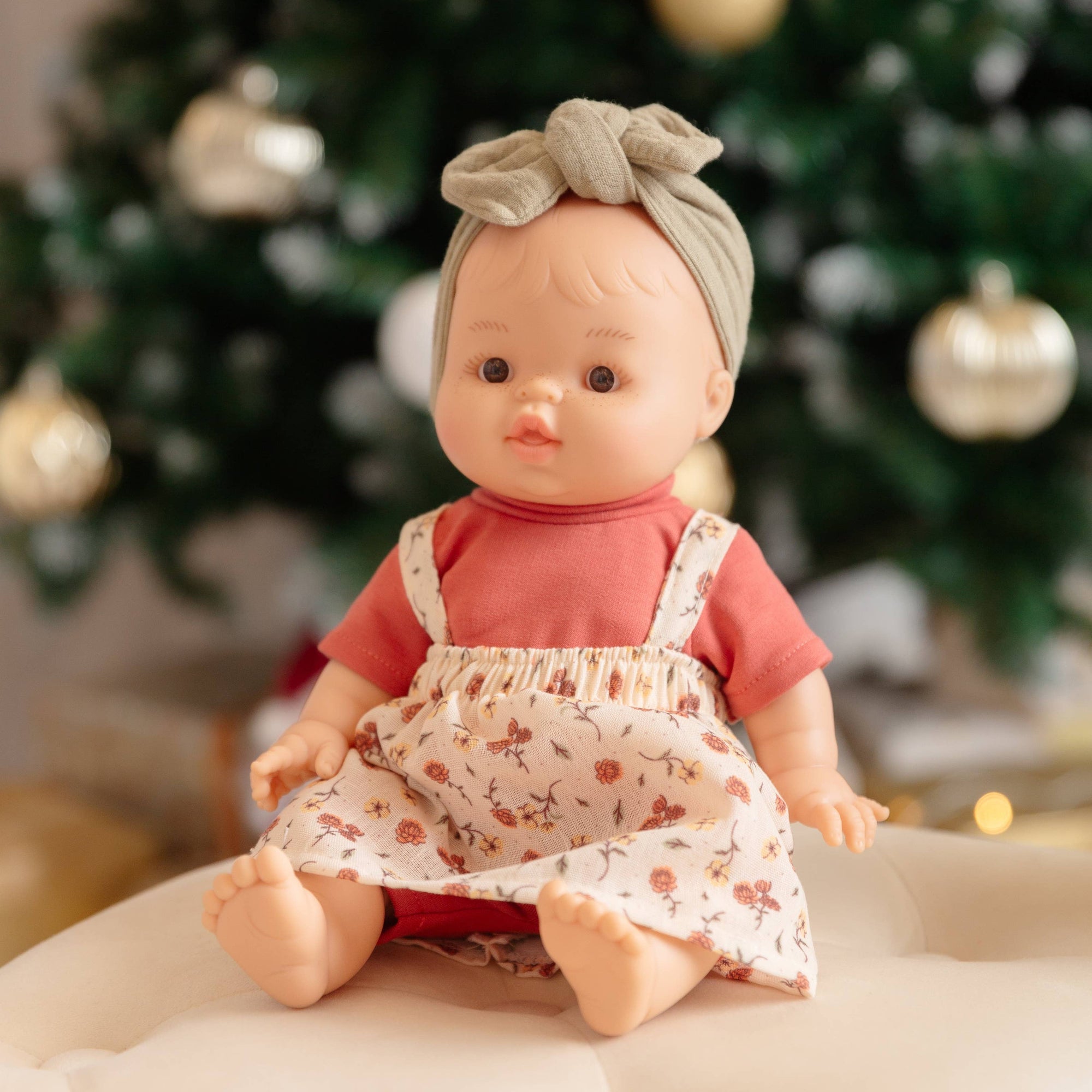 Doll in a floral dress sitting in front of a decorated Christmas tree.
