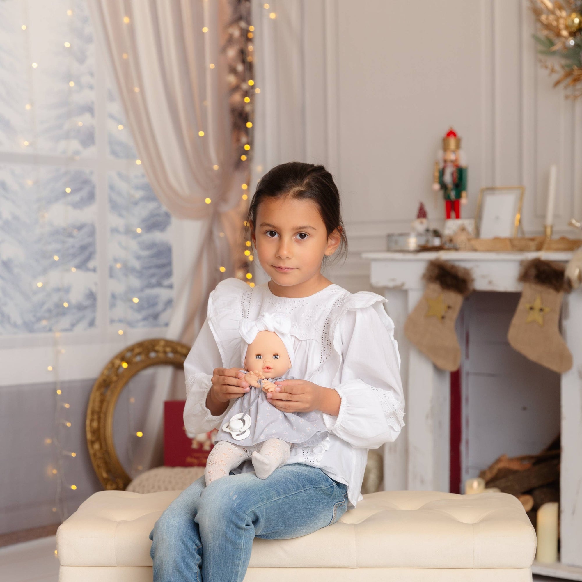 Child sitting on a stool holding a baby doll in a decorated room with lights and stockings.