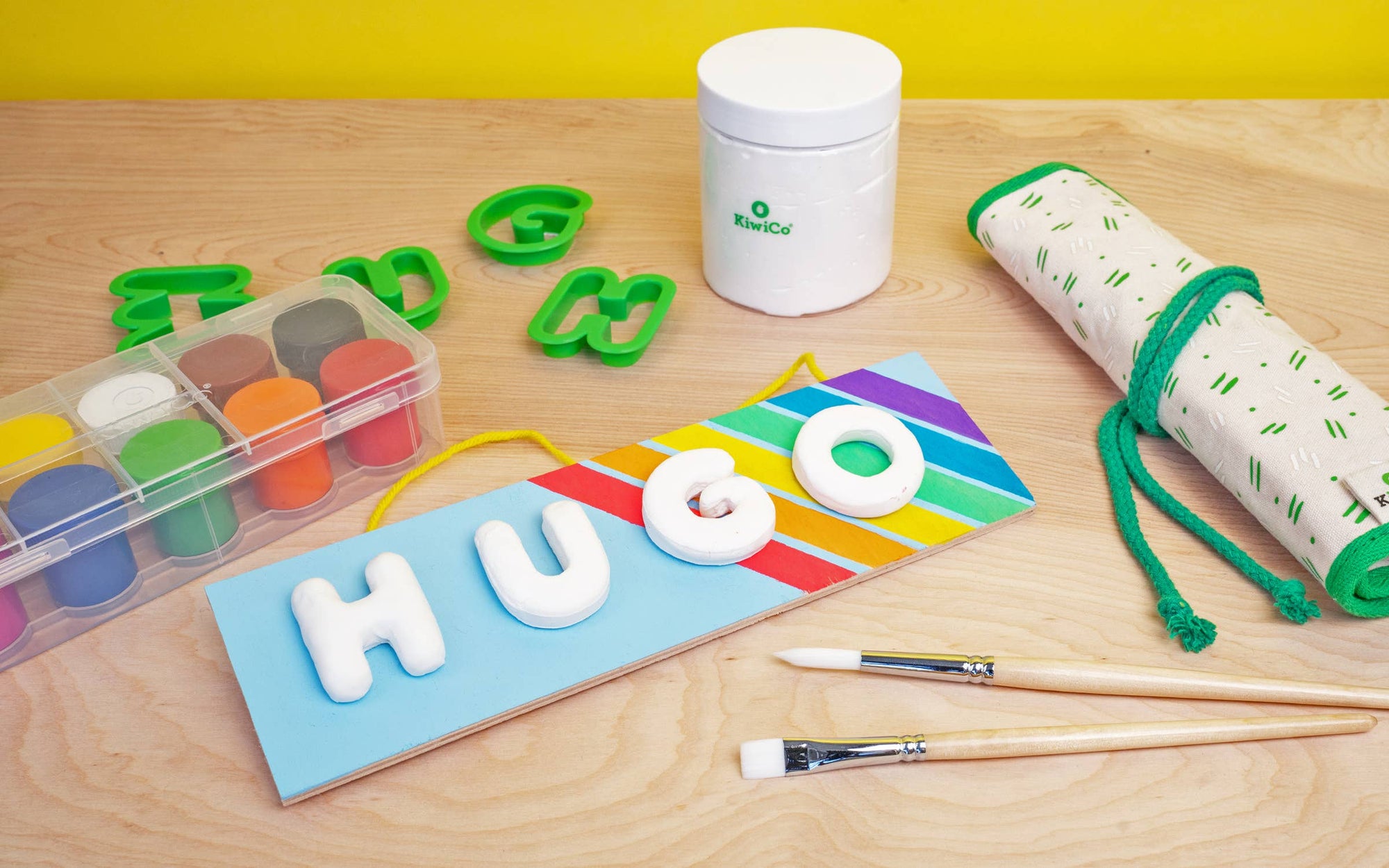 A children's craft kit laid out on a light wood surface with a bright yellow backdrop. The kit includes a white container labeled BioToc, green letter cutters, a clear palette of paint colors, a rolled cream-and-green fabric item, two paintbrushes, and a blue board with colorful diagonal stripes and white letters spelling HUGO.