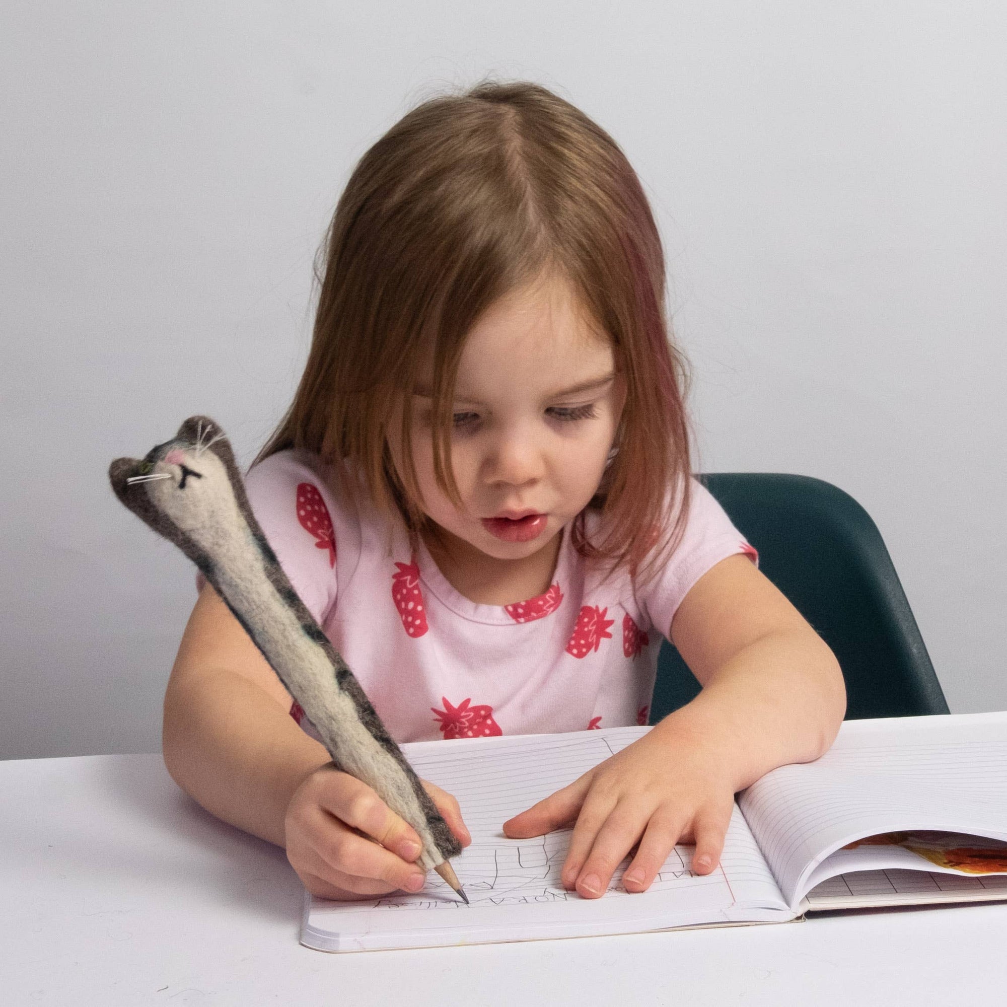 Child writing with a stylus on a book, wearing a pink shirt with red patterns.