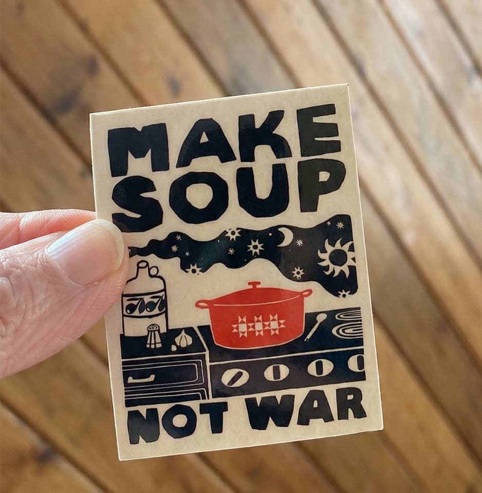 Sticker with 'Make Soup Not War' text held by a hand on a wooden surface