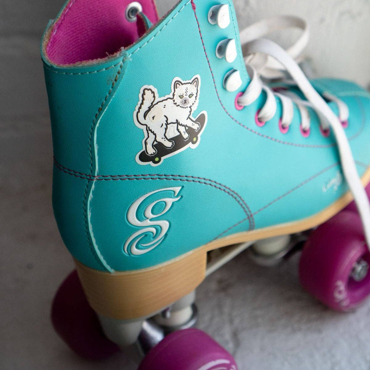 Turquoise roller skates with pink accents and a cat illustration on a neutral background