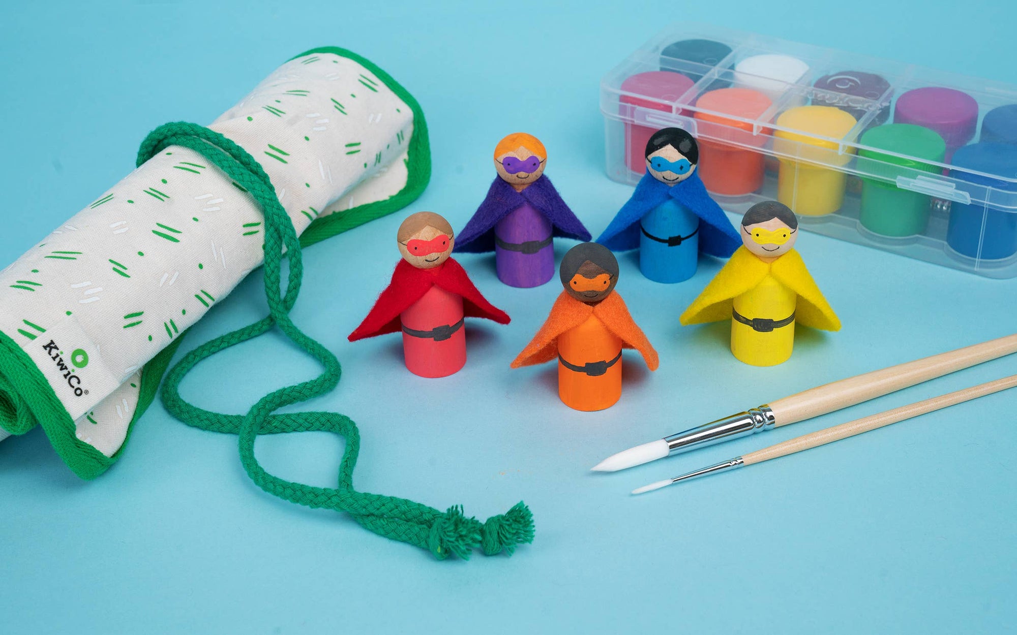 A children's painting kit with five unpainted wooden superhero figures in red, orange, blue, yellow, and purple, a green drawstring bag, a clear paint palette, and two paintbrushes on a light blue background.