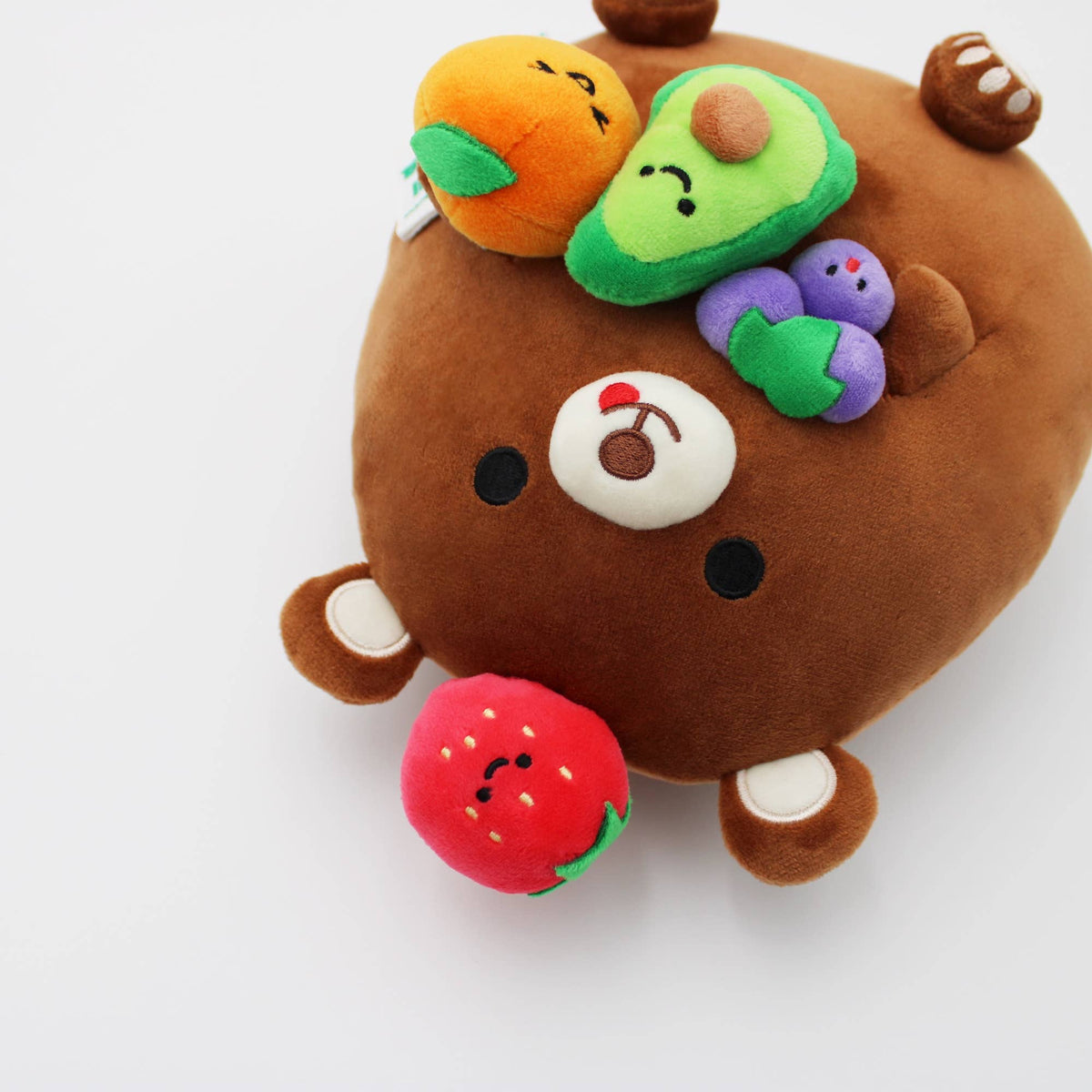 A brown plush bear toy with embroidered facial features and several colorful fruit-shaped accessories attached to its head and body, photographed on a clean white background.