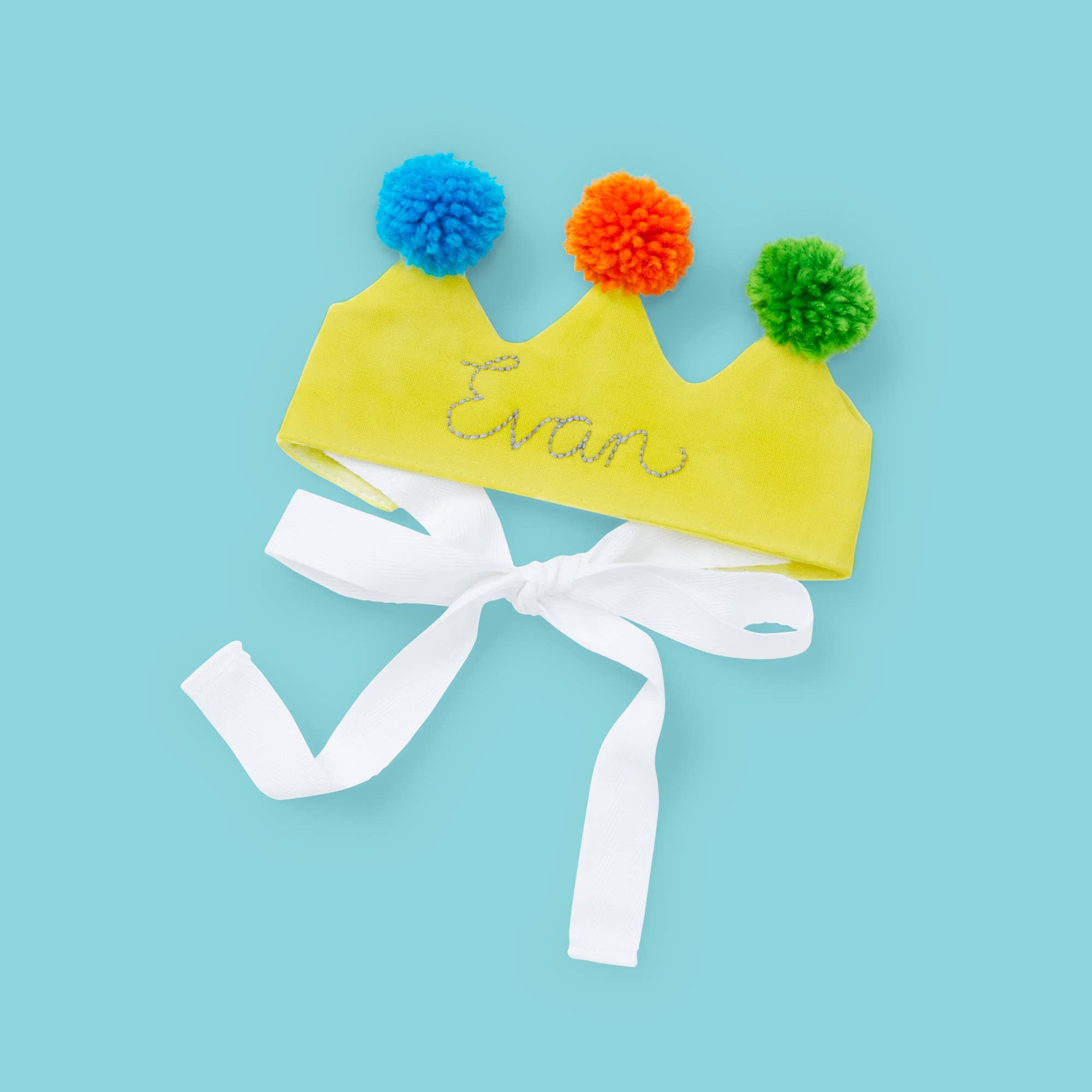 A yellow fabric crown costume accessory with three colorful pom-poms in blue, orange, and green, personalized with the name 'Evan' in silver glitter, and finished with a white ribbon bow, shown on a light blue background.