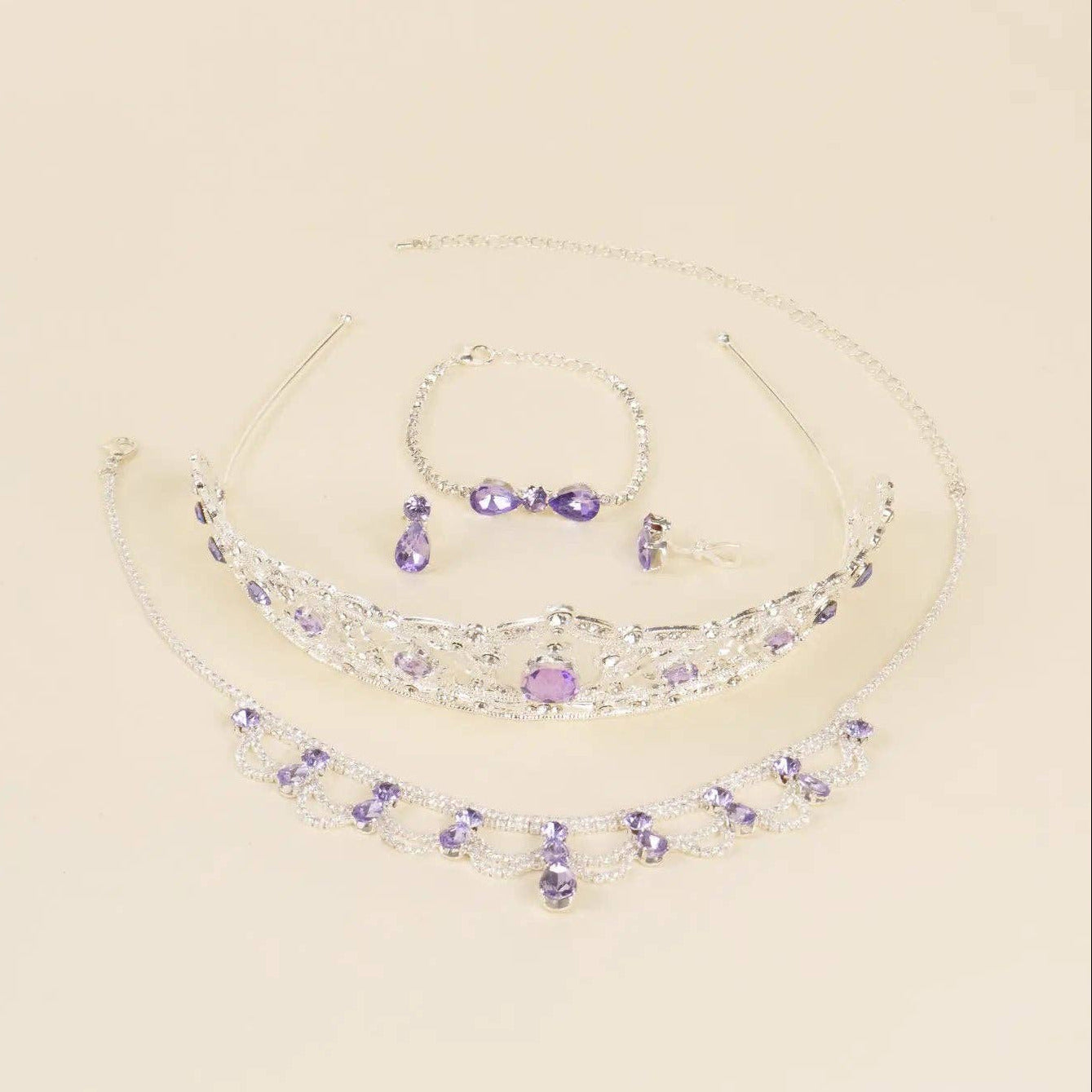 A silver-toned jewelry set featuring a tiara, necklace, bracelet, and earrings, embellished with clear rhinestones and purple gemstones, displayed on a light beige background.