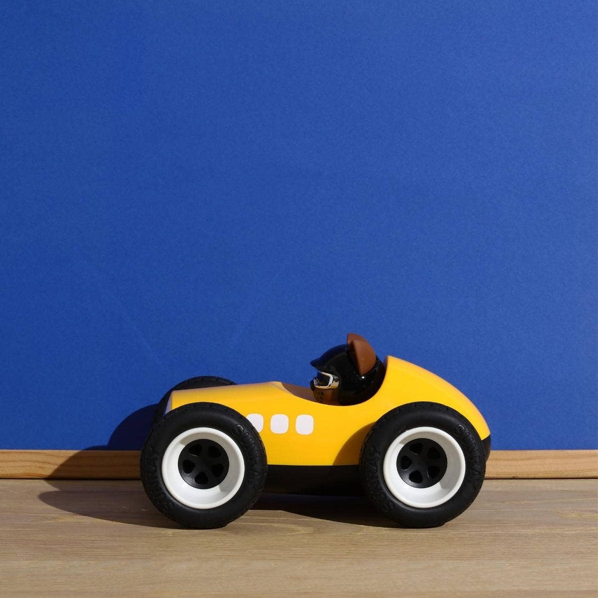 A small yellow toy race car with black wheels and white rims, featuring a cartoonish face on the front, positioned on a light wood floor against a solid blue wall.