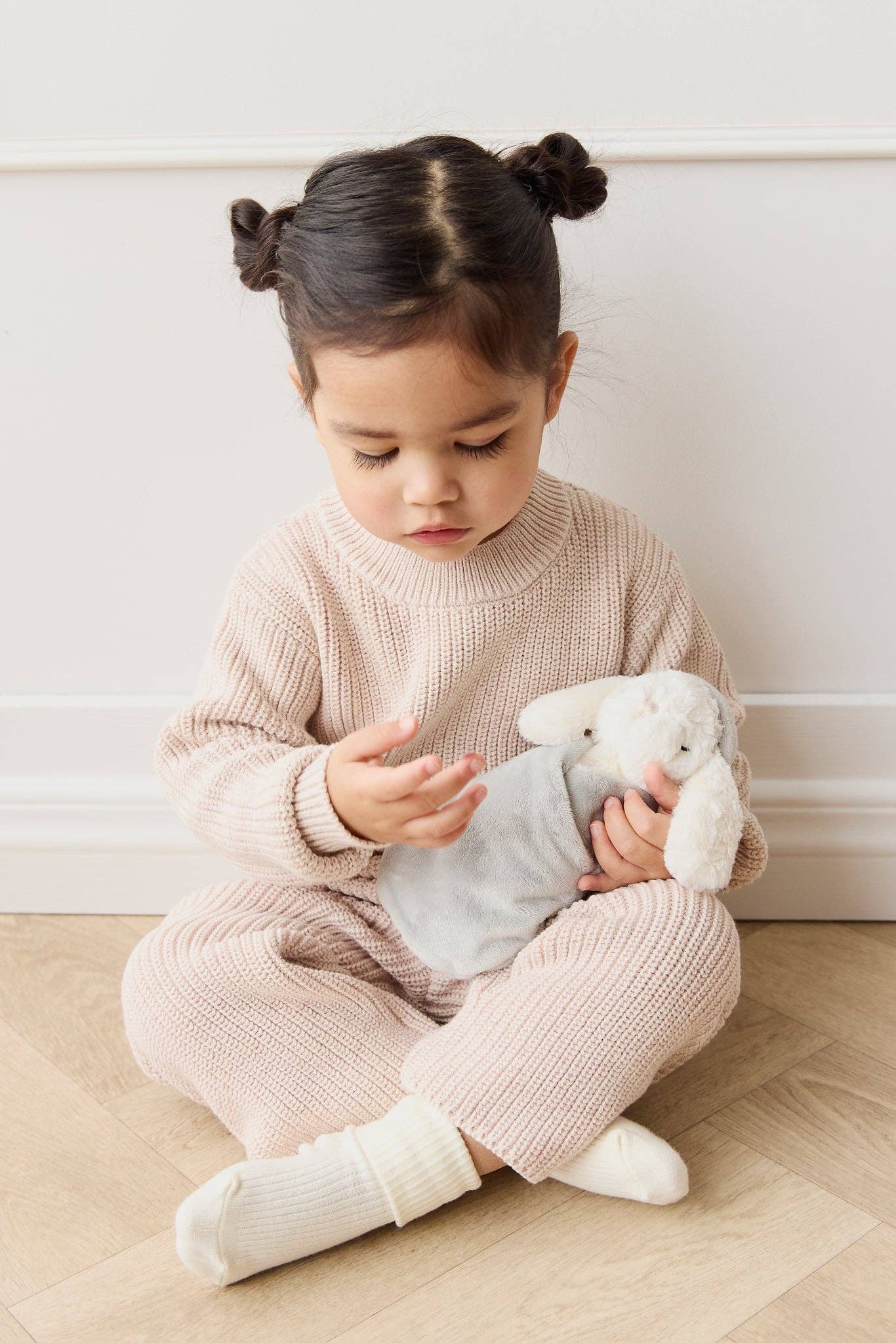 Child sitting on the floor holding a teddy bear against a white wall.