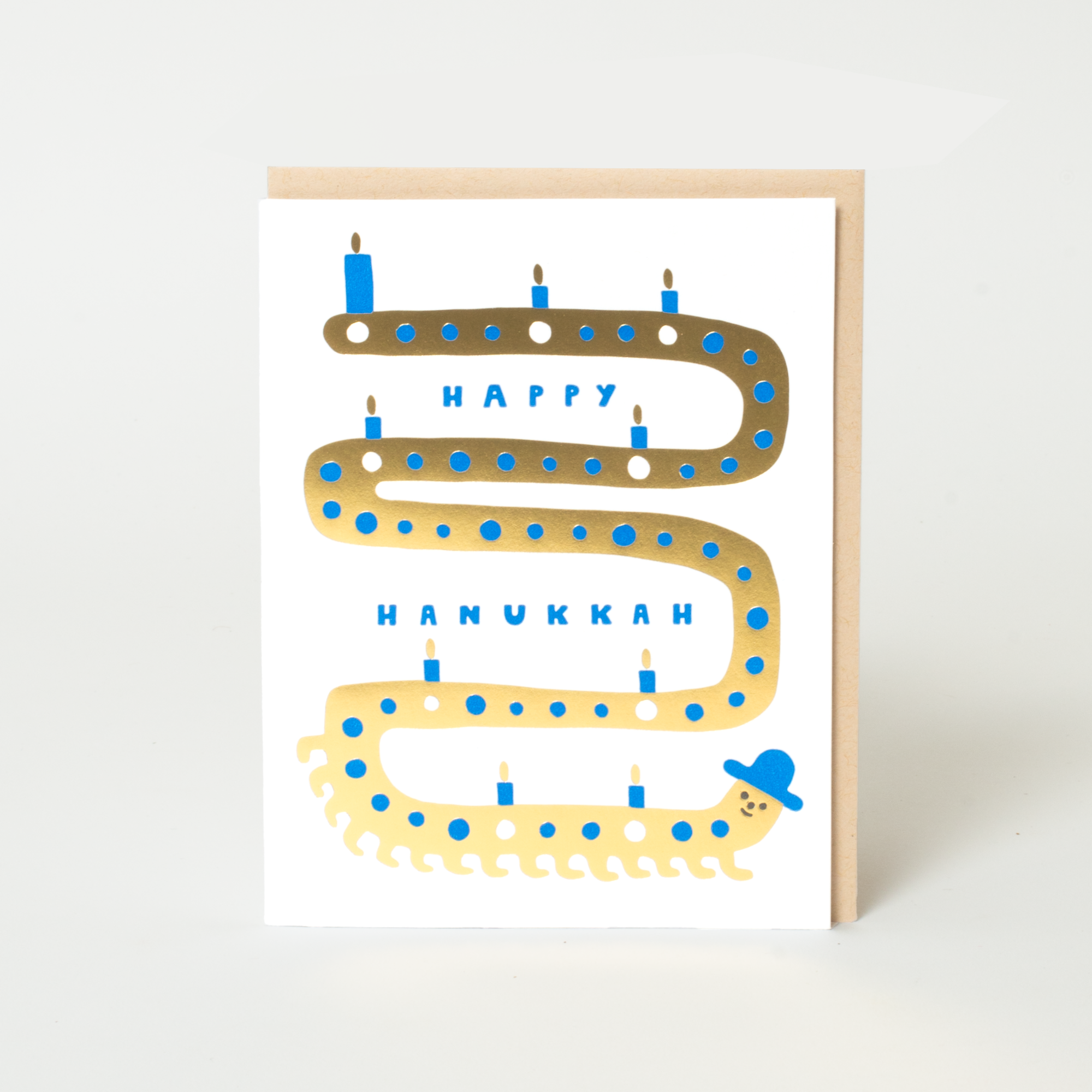 A white Hanukkah greeting card with a gold and blue illustrated serpentine candle path, the words 'HAPPY HANUKKAH,' and a small cartoon figure wearing a blue hat, shown with a light wood envelope on a white background.