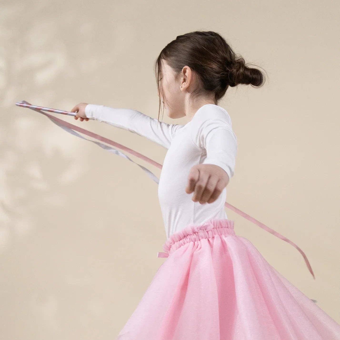 A young girl in a white long-sleeve top and a fluffy pink tutu skirt twirling with a pink ribbon against a warm beige background.