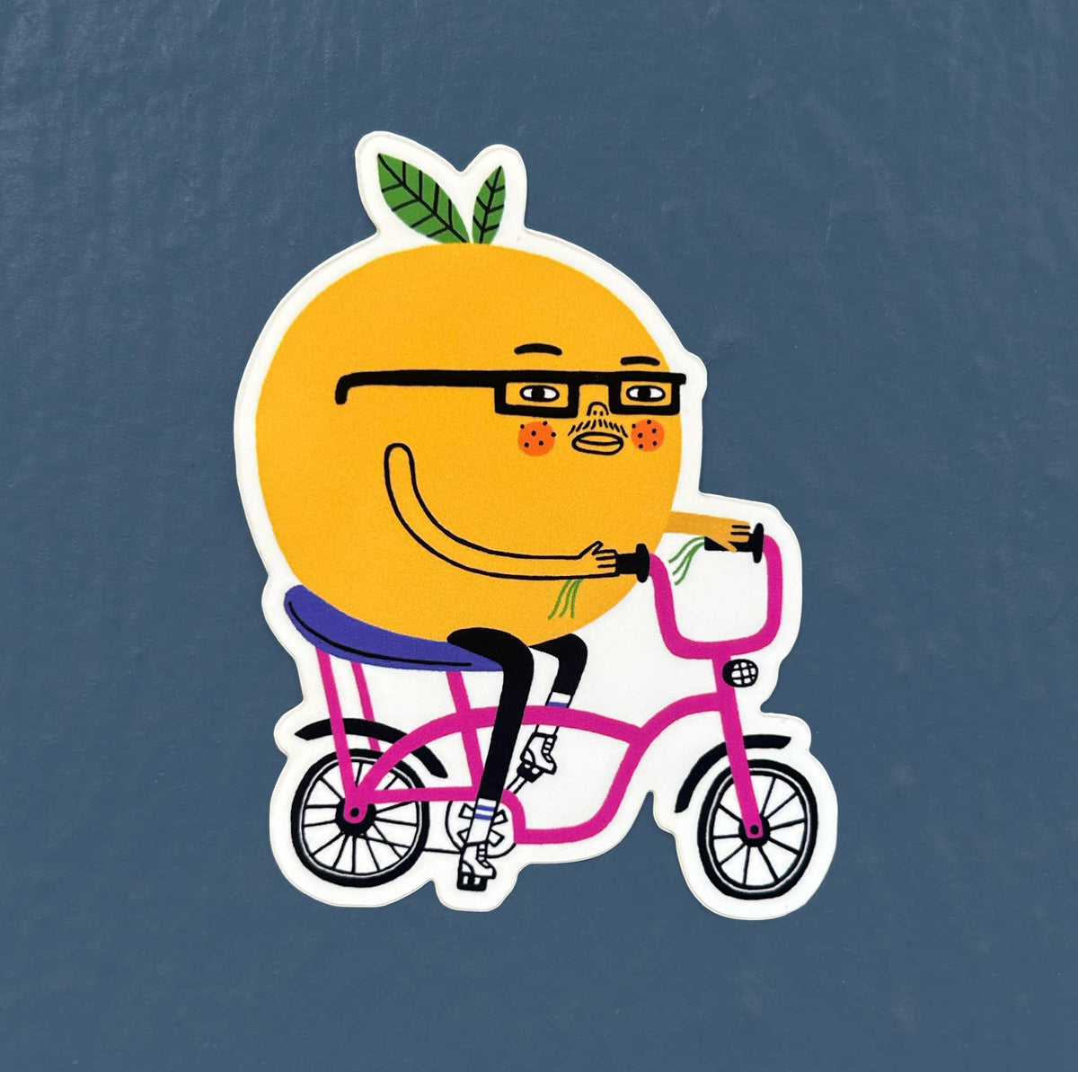 A die-cut cartoon sticker of a yellow citrus fruit character wearing black glasses and riding a pink bicycle, shown on a textured dark slate blue background.
