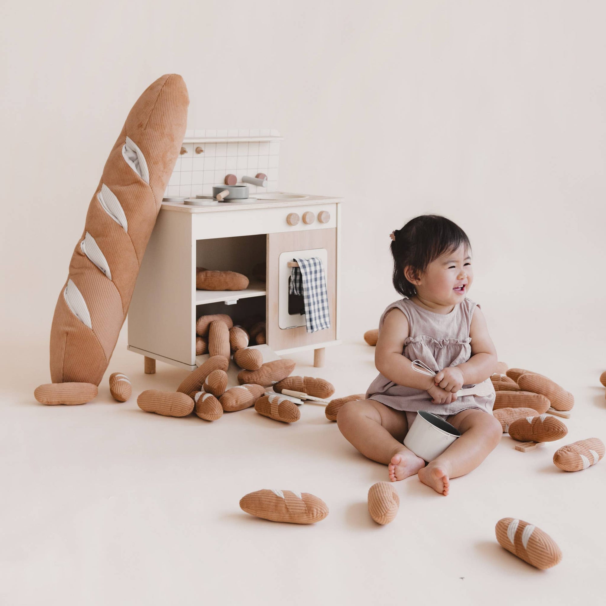 Child playing with toy bread and a small wooden kitchen set on a light-colored floor.
