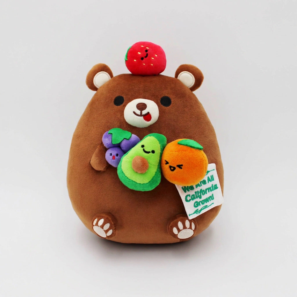 A brown plush bear toy with cute fruit accents, including a strawberry on its head, holding an avocado and an orange, with a small tag attached, photographed on a clean white background.
