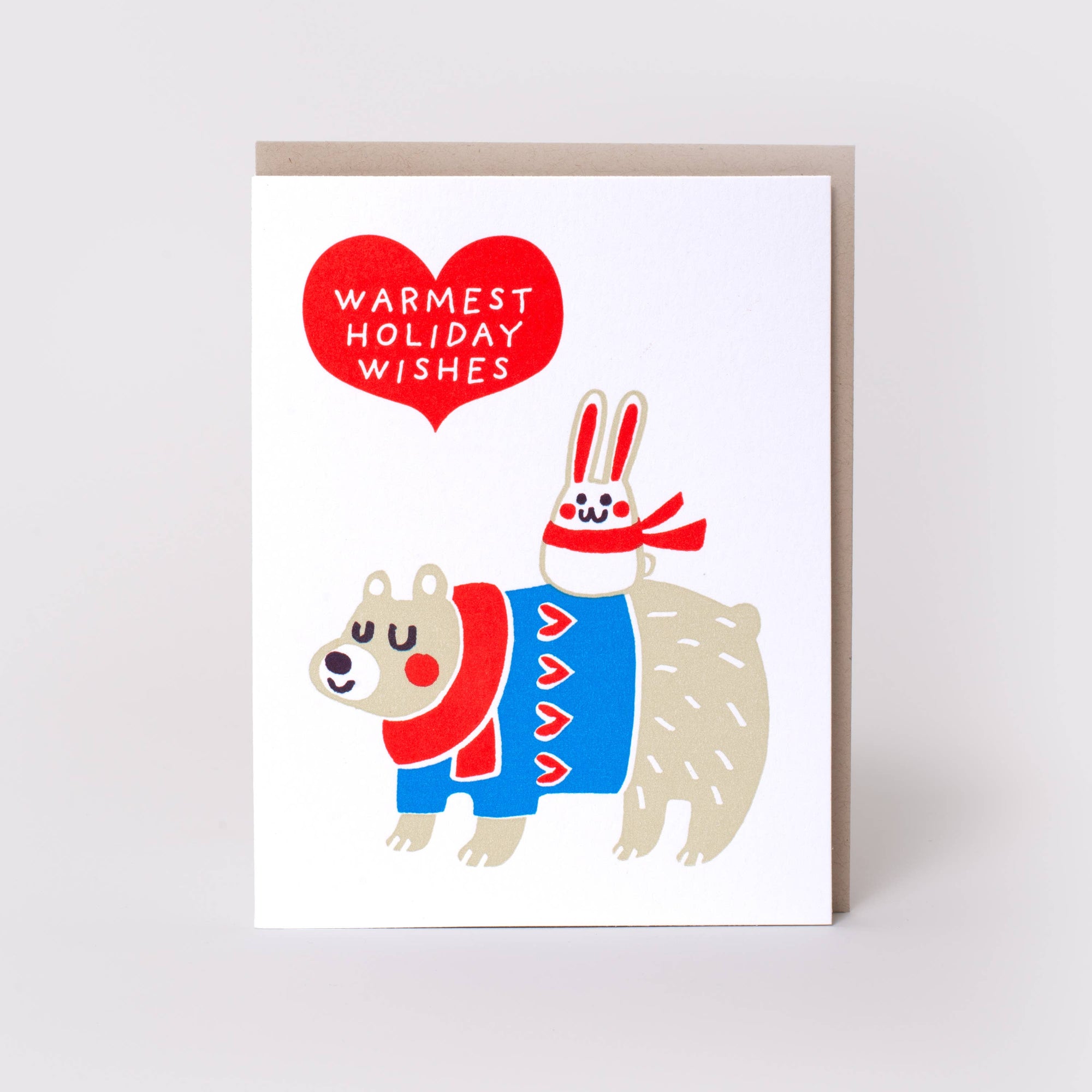 A greeting card with a white front featuring a cartoon polar bear and rabbit in red and blue winter clothing, with a red heart that reads 'WARMEST HOLIDAY WISHES.' A kraft envelope is visible behind the card on a light gray background.