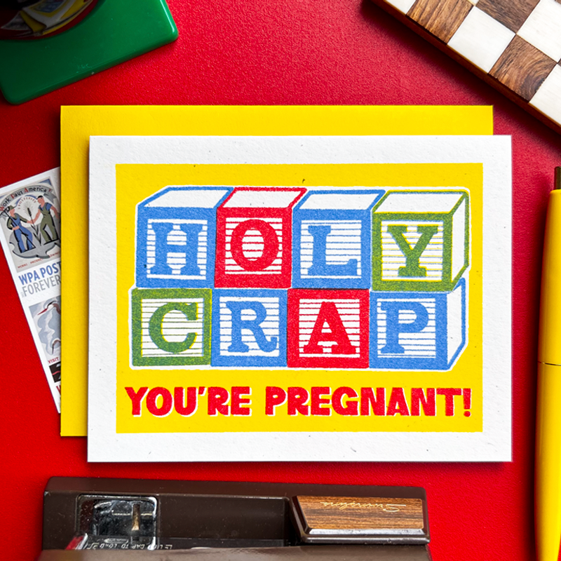 A novelty greeting card with a bright yellow border and a playful block-letter design spelling 'HOLY CRAP' above the words 'YOU'RE PREGNANT!'. The card is photographed on a red surface with a few surrounding props, including a yellow card underneath, a pen, a stamp, and a checkerboard corner.