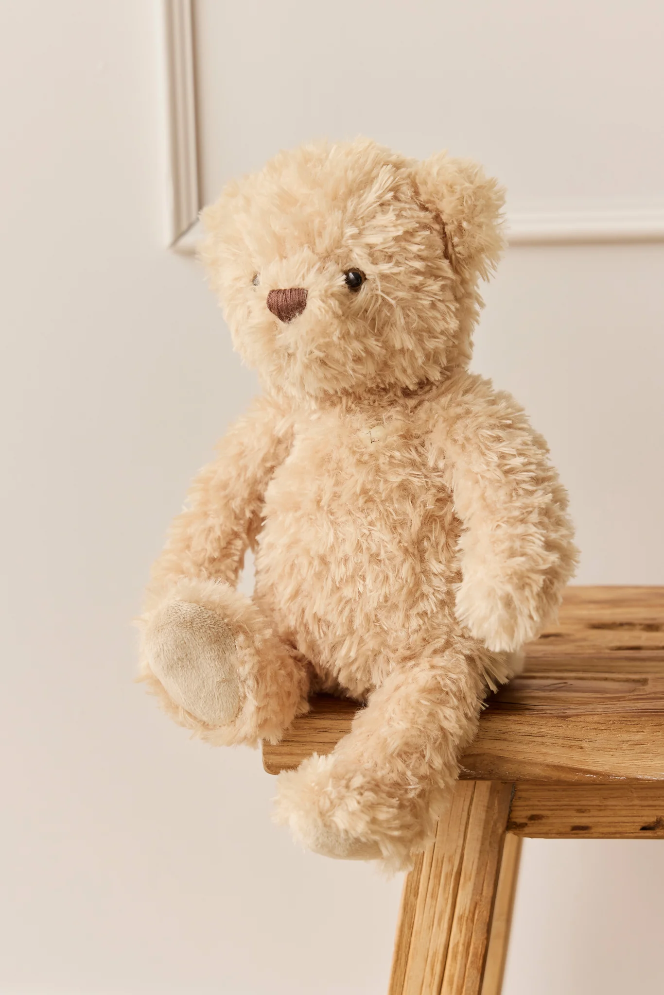 Beige teddy bear sitting on a wooden stool against a light background