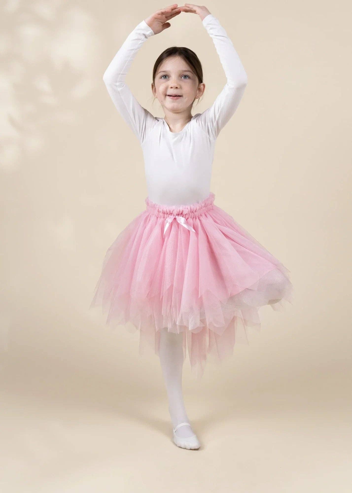 A young girl dancer posing in a white long-sleeve leotard and a fluffy light pink tutu skirt with white tights, set against a warm beige studio backdrop.