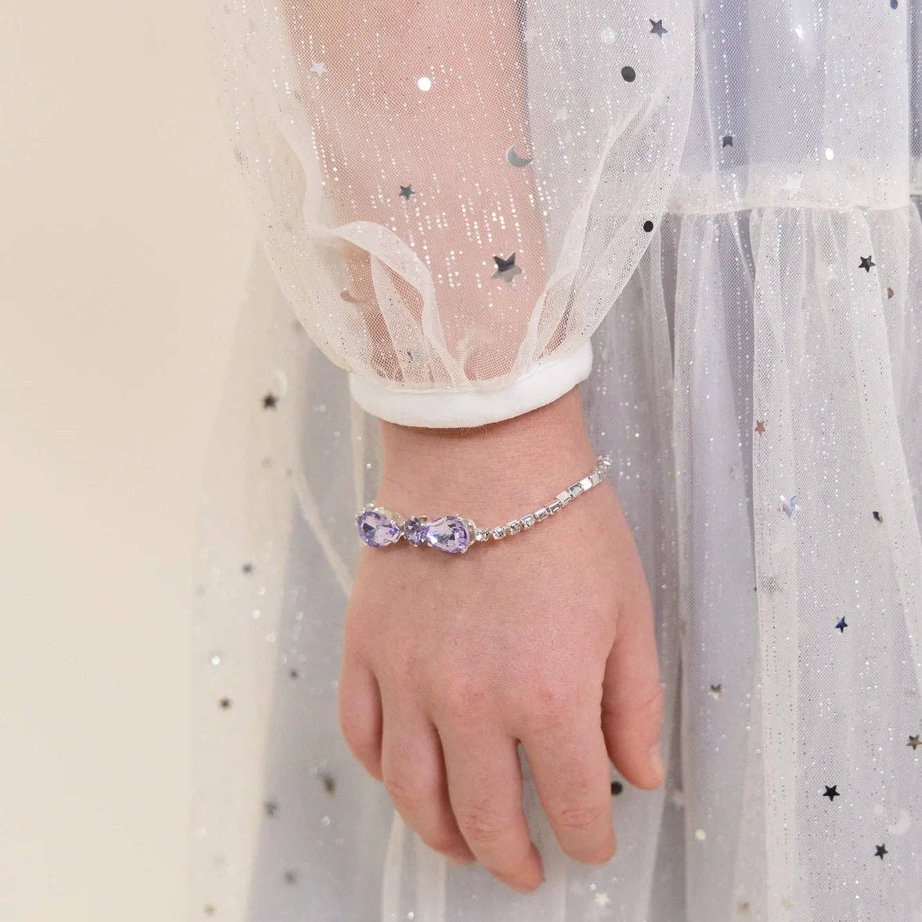 A close-up of a child's hand wearing a silver-toned chain bracelet with a purple bow-shaped crystal centerpiece, styled with a sparkly white tulle dress.