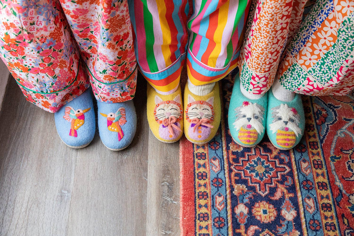 Four pairs of colorful, patterned knit slipper socks worn by different people, shown from the knees down on a wooden floor and patterned rug.