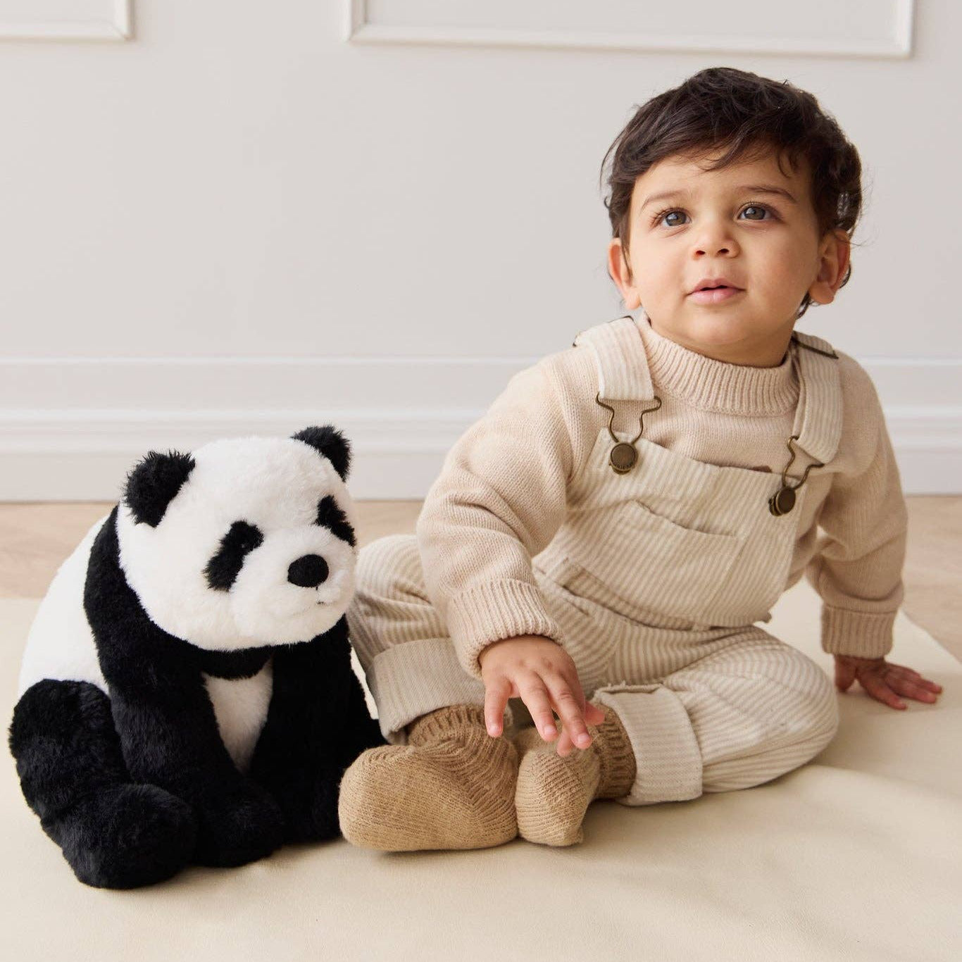 Child in a beige outfit sitting next to a plush panda toy on a light-colored floor.