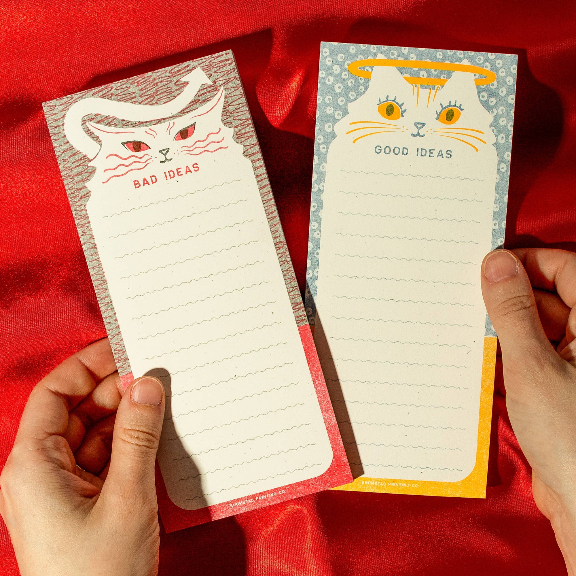 Two notepads with cat illustrations and text on a red fabric background