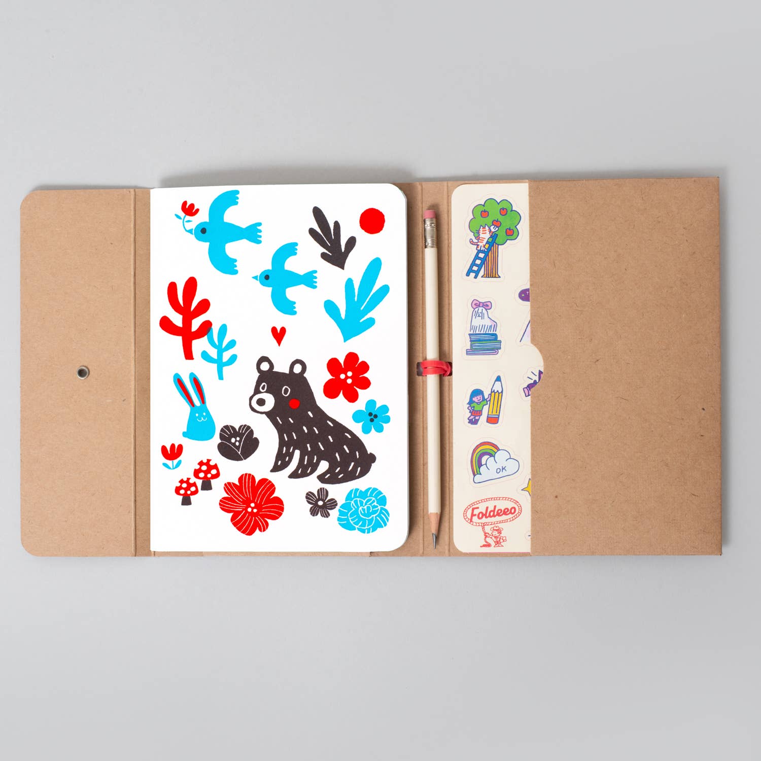 A kraft paper notebook shown open on a light gray background, featuring a white sticker sheet with whimsical illustrations of a bear, birds, flowers, and leaves, alongside a pencil and a sticker strip pocket.