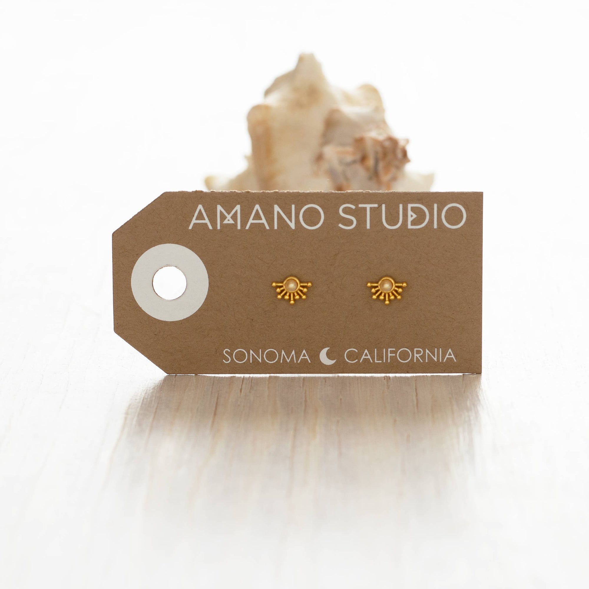 Gold earrings on a 'Amano Studio' branded card with a shell in the background
