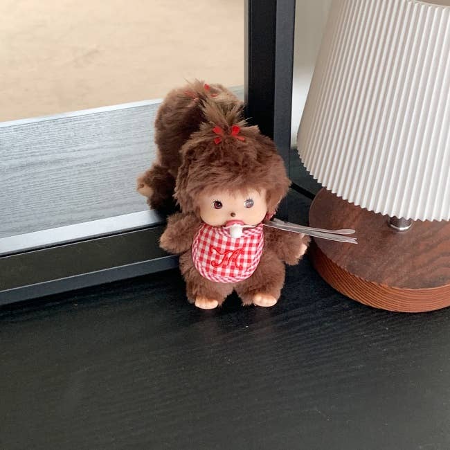 Small monkey plush toy with a checkered dress on a dark surface next to a lamp.