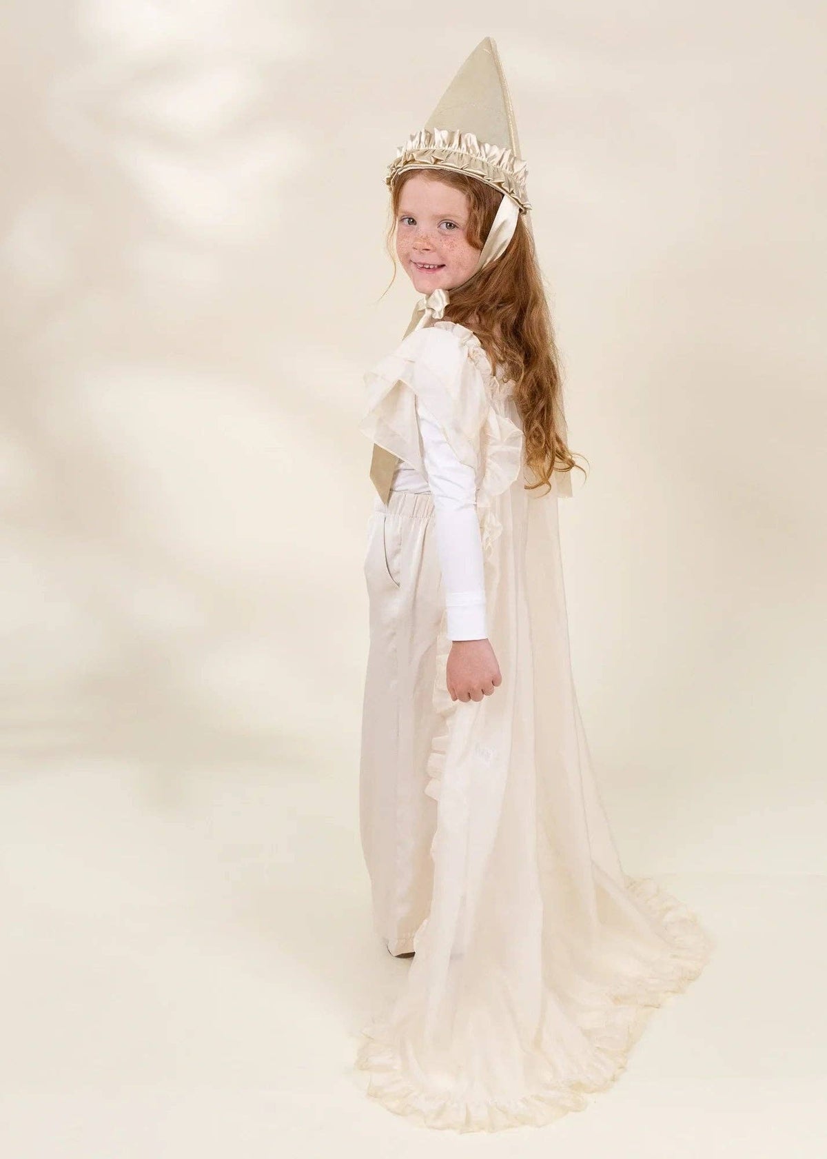 A young girl with long light brown hair models a white and beige fairy or princess costume, featuring a long-sleeved dress, a matching pointed hat with a jeweled crown, and a sheer cape, set against a warm cream-colored studio background.