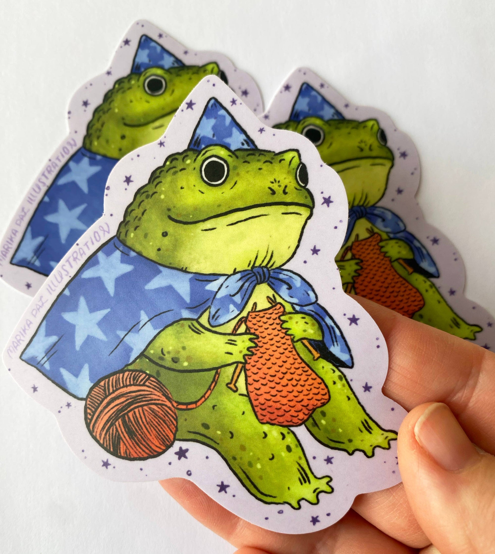 Hand holding a card with a green frog character holding yarn and knitting needles on a white background