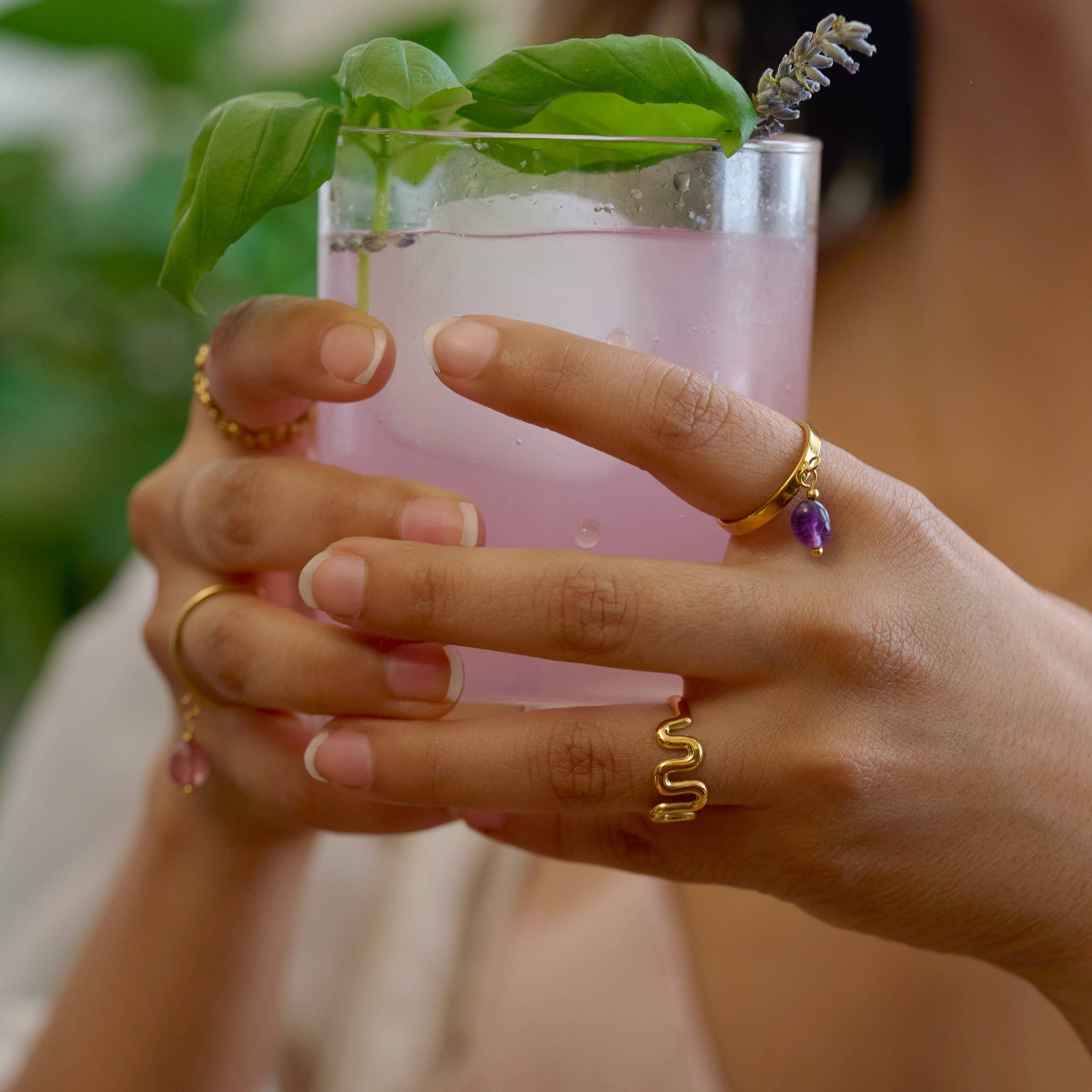 Person holding a pink cocktail with a sprig of mint, wearing gold rings.