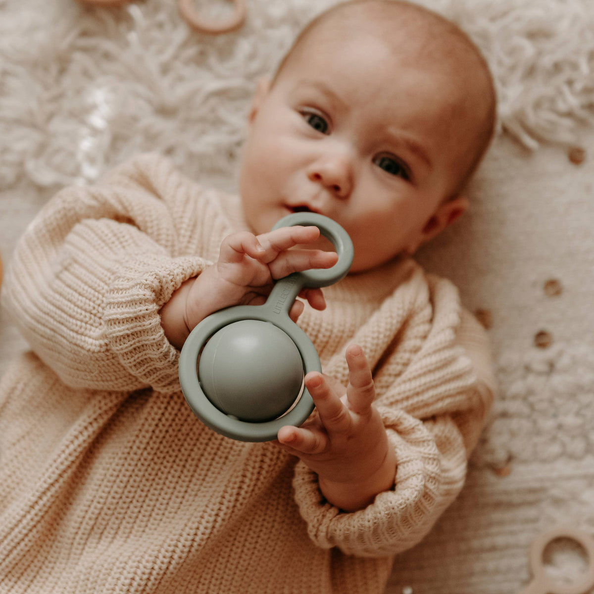 Baby lying on a textured surface with a teething ring and wooden toys around.